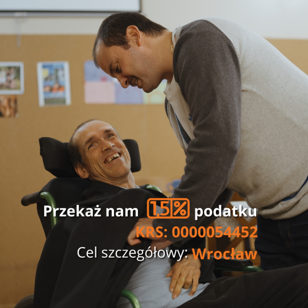 Zdjęcie przedstawia dwóch mężczyzn w ciepłej i radosnej interakcji. Jeden z nich, mężczyzna o szczupłej twarzy i krótkich, ciemnych włosach, siedzi na wózku inwalidzkim. Ma na sobie czarną bluzę i szeroko się uśmiecha, patrząc na drugiego mężczyznę. Drugi mężczyzna, stojący obok, pochyla się w jego stronę z serdecznym uśmiechem. Ma krótkie, ciemne włosy i nosi szaro-białą bluzę z długim rękawem. Ich dłonie są blisko siebie, co sugeruje bliskość i troskę. Tło to tablica korkowa z przypiętymi kolorowymi zdjęciami i rysunkami, co sugeruje domową lub ośrodkową atmosferę. Na zdjęciu znajduje się tekst w języku polskim: "Przekaż nam 1,5% podatku", napisany białą czcionką, z wyróżnionym na pomarańczowo numerem KRS: 0000054452. Poniżej widnieje informacja o celu szczegółowym: "Wrocław", również podkreślona pomarańczowym kolorem. Przekaz zdjęcia jest pozytywny – przedstawia przyjaźń i wsparcie dla osób z niepełnosprawnością, jednocześnie zachęcając do przekazania 1,5% podatku na rzecz organizacji pomagającej takim osobom.