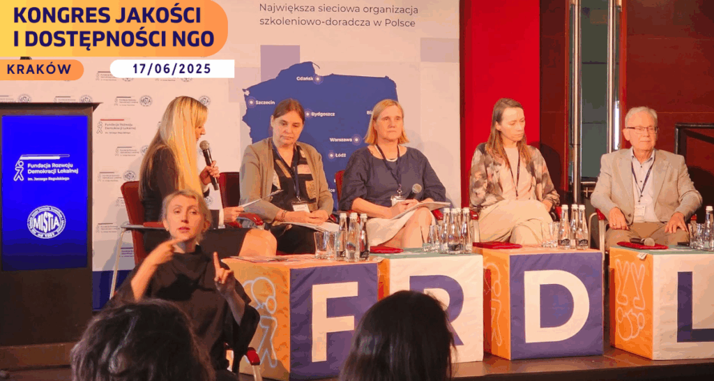 Na zdjęciu widać scenę z pięcioosobowym panelem dyskusyjnym. Paneliści siedzą na krzesłach ustawionych w rzędzie. Przed nimi stoją kolorowe kostki z literami tworzącymi skrót „FRDL”, nawiązujący do Fundacji Rozwoju Demokracji Lokalnej. Kostki są w kolorach pomarańczowym, granatowym i niebieskim, z graficznymi piktogramami związanymi z fundacją. Od lewej strony siedzi kobieta z długimi blond włosami, trzyma mikrofon i prowadzi rozmowę. Obok niej — trzy kobiety i jeden mężczyzna, wszyscy mają identyfikatory na smyczach. Wszyscy są ubrani w półformalny sposób, siedzą przy stole z butelkami wody i szklankami. W lewym dolnym rogu zdjęcia widoczna jest tłumaczka Polskiego Języka Migowego w czarnej bluzce, wykonująca gesty w kierunku publiczności. Tło stanowi niebiesko-biała plansza z logotypem Fundacji Rozwoju Demokracji Lokalnej im. Jerzego Regulskiego. Widnieje na niej mapa Polski z zaznaczonymi miastami, w których znajdują się oddziały fundacji. Nad mapą widnieje hasło: „Największa sieciowa organizacja szkoleniowo-doradcza w Polsce”. W górnym rogu zdjęcia, po prawej stronie, znajduje się pomarańczowo-fioletowy baner z napisem: „Kongres Jakości i Dostępności NGO, Kraków, 17/06/2025”. Na sali obecni są uczestnicy, których sylwetki częściowo widać od tyłu, na pierwszym planie.