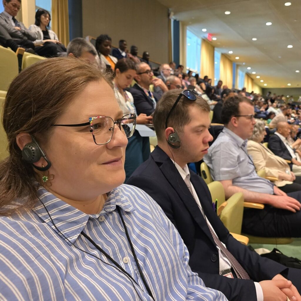 Sala konferencyjna wypełniona uczestnikami siedzącymi w zielonych fotelach. W pierwszych rzędach widać osoby w słuchawkach do tłumaczeń, a dalej wielu słuchaczy notujących lub śledzących wystąpienie. Jasne, przestronne wnętrze i formalny charakter spotkania podkreślają międzynarodowy, profesjonalny kontekst wydarzenia
